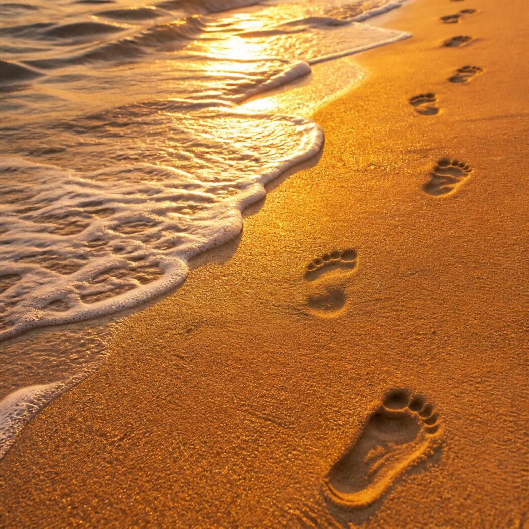 Träume, die Flügel haben 3 In Grossaufnahme goldfarbener Sand ueber den lebhafte Wellen des Meeres spuelen Fussspuren fuehren aus dem Wasser ans Land das Bild ist vom Goldlicht durchflutet realistisch magisch